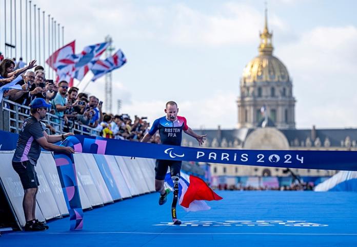 A male Para triathlon athlete with a prosthetic running blade is crossing the finish line in front of a packed crowd with the Invalides in the background